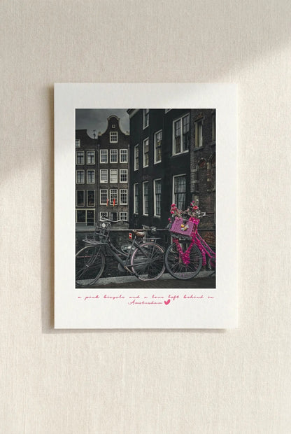 Amsterdam postcard featuring a vintage pink bicycle beside canal houses on a beige wall. 